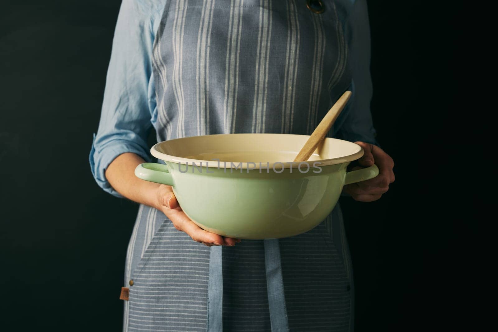 Crop anonymous female cook in apron holding bowl with spatula while preparing for baking against black background in modern studio