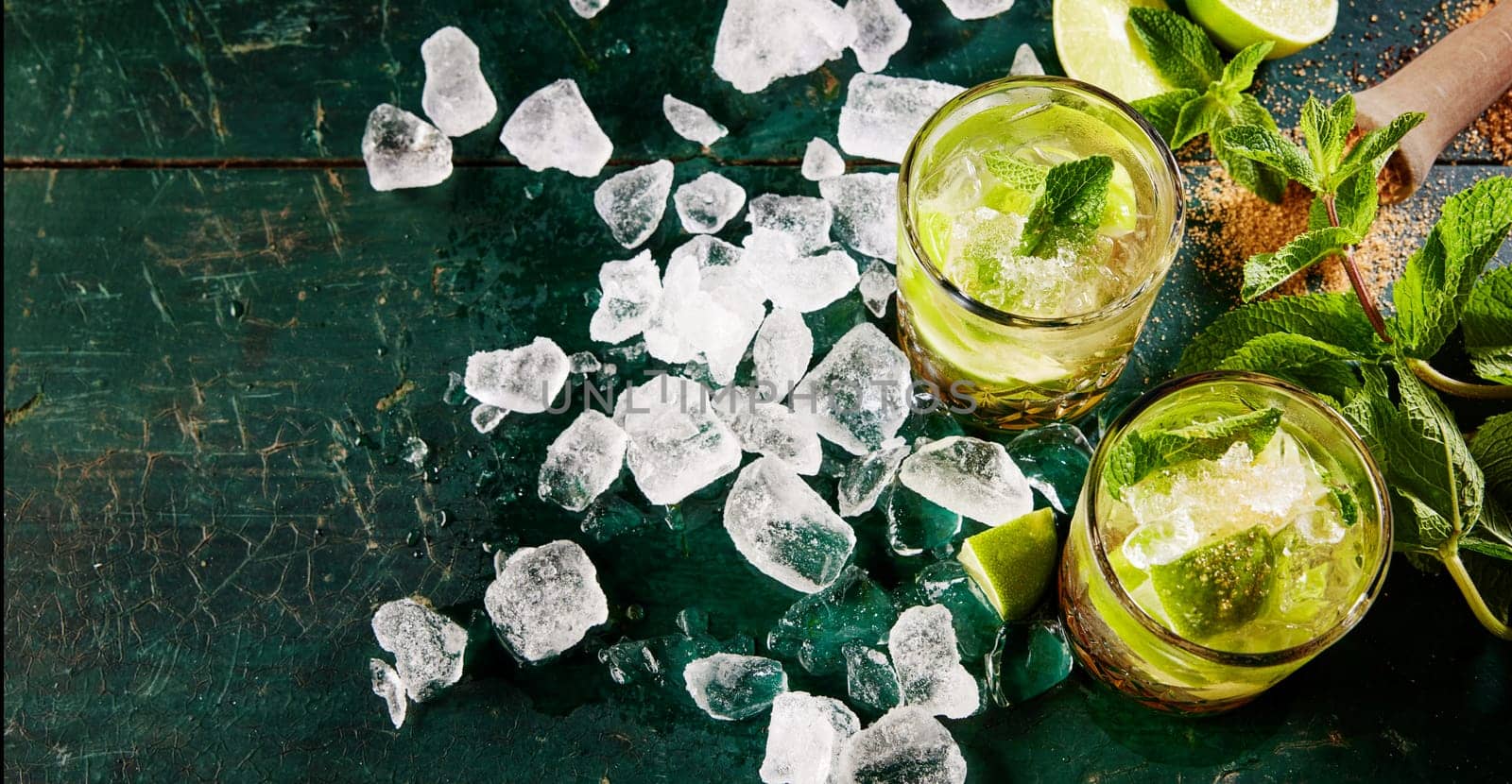 Top view of tasty alcoholic mojito beverages served on shabby dark table with scattered ice cubes and mint leaves in kitchen