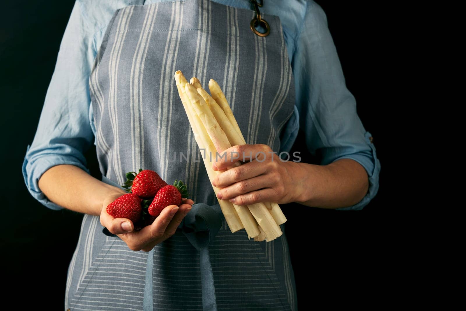 Crop anonymous female cook in apron holding bunch of white celery and ripe red strawberries against black background during cooking preparation