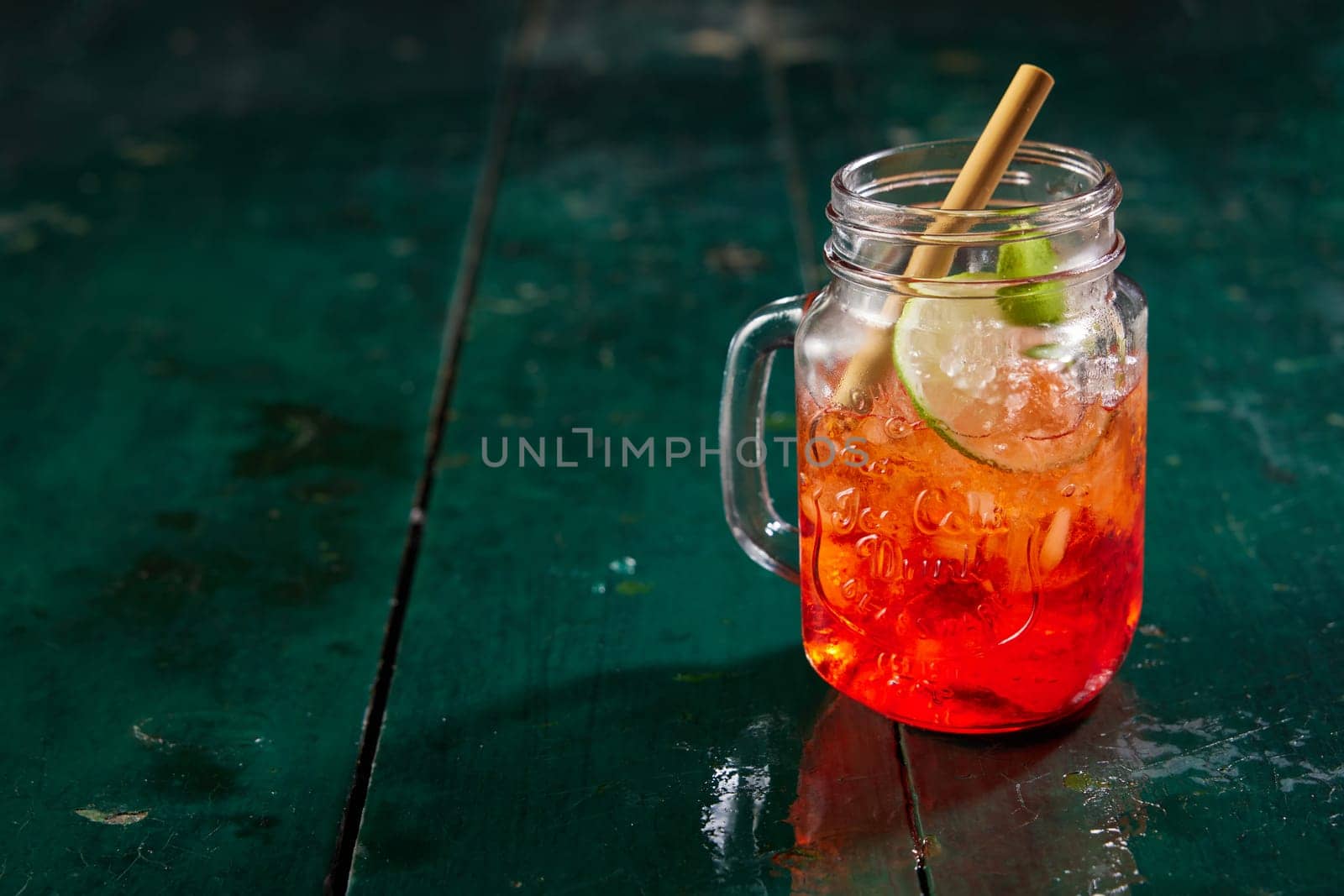 Glass jar filled with refreshing sweet red berry beverage served with lime and straw on dark wooden table