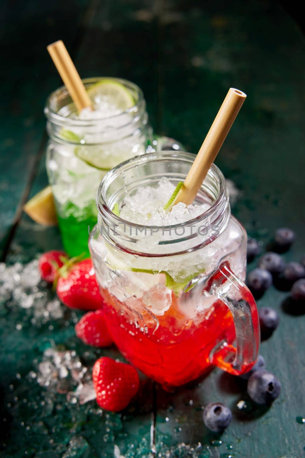 From above of glass jars filled with red and green berry beverages served with straws on table with strawberries and blueberries in kitchen
