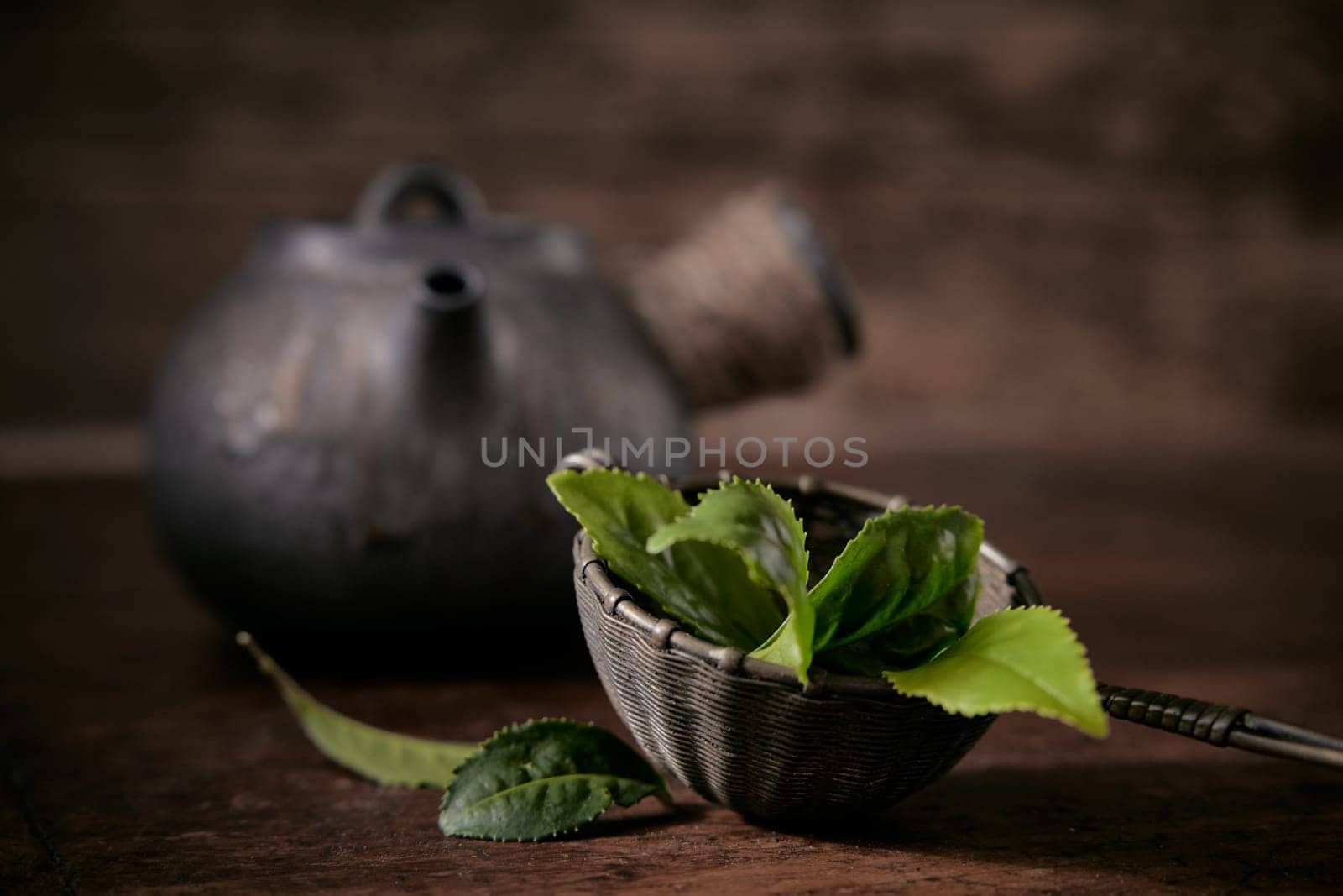 Fresh green tea leaves in ladle placed on table near old fashioned metal teapot on blurred background in light room