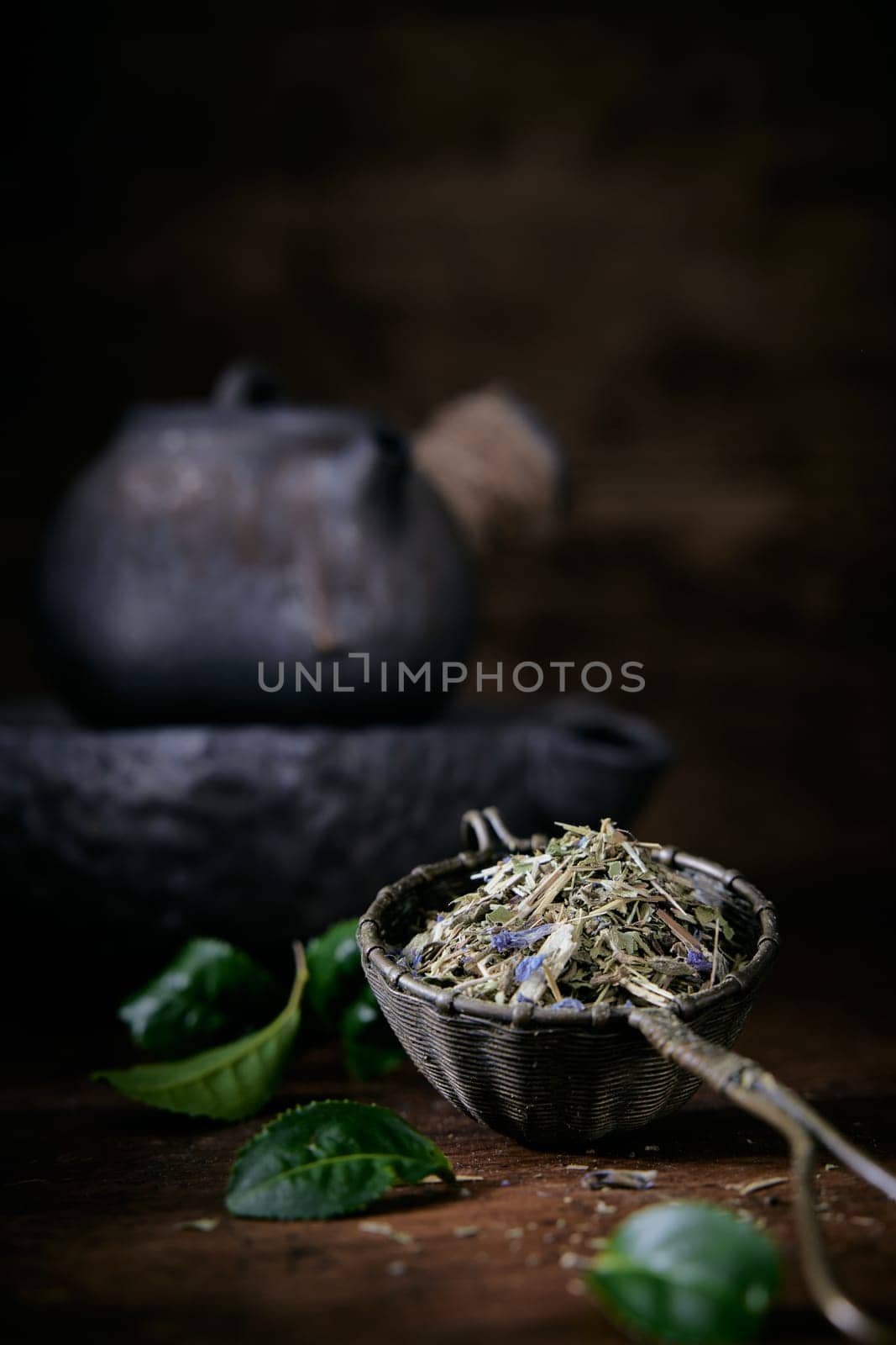 Aromatic loose tea and fresh green leaves placed on table with old fashioned metal teapot on blurred background in room