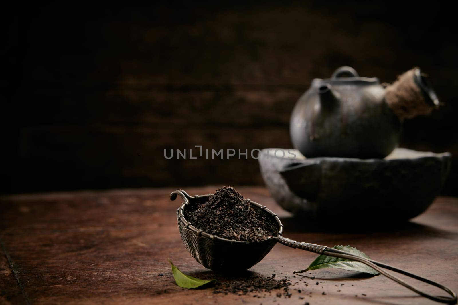 Pile of aromatic loose tea for brewing placed on wooden table with fresh green leaves and old teapot in kitchen