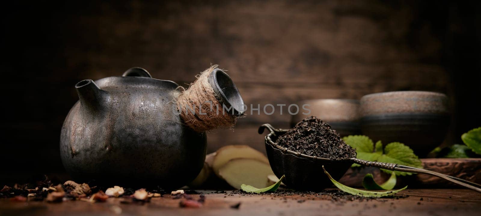 Aromatic loose tea near old fashioned teapot placed on table with scattered condiments and ginger in kitchen with set of cups