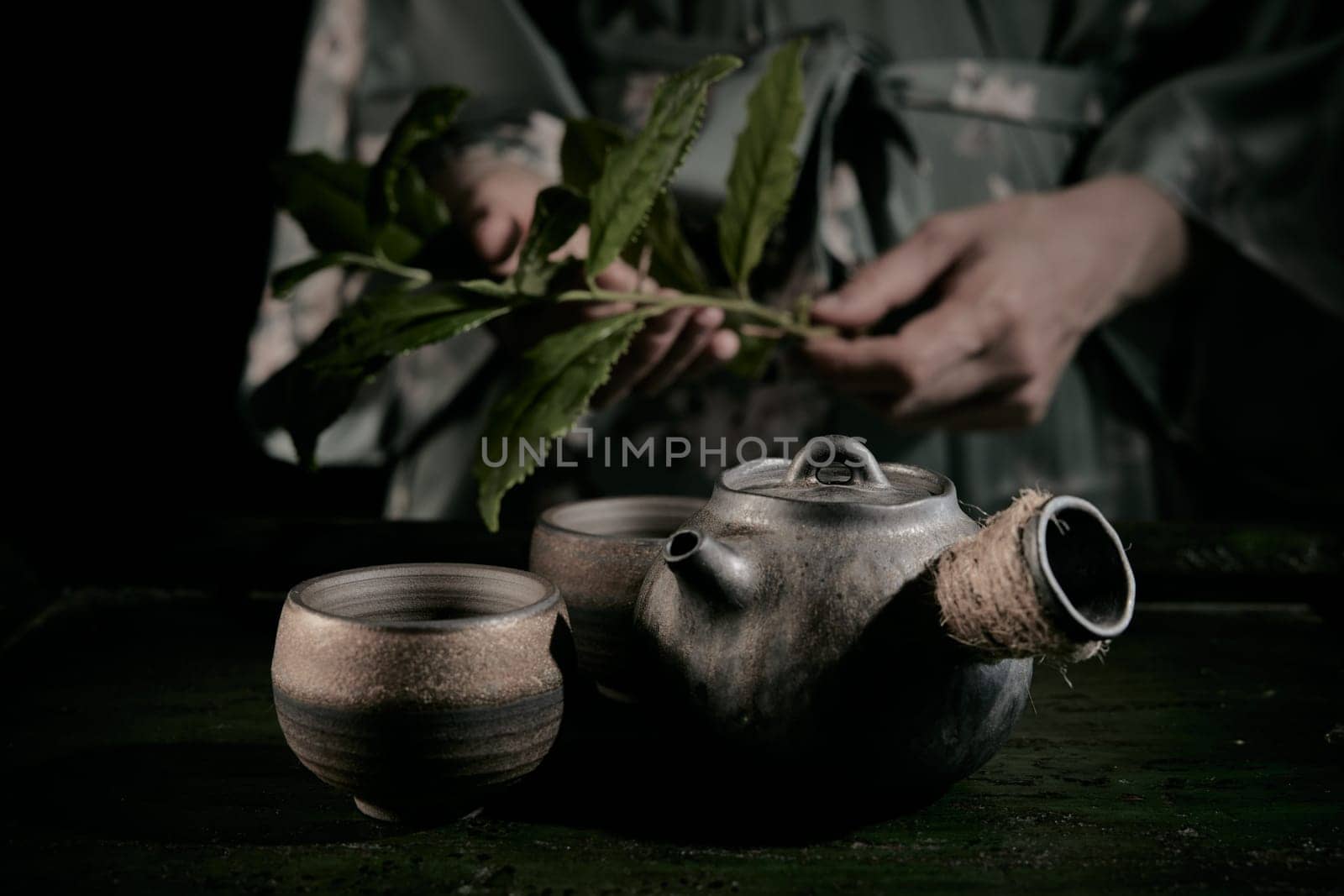 Crop unrecognizable person with fresh green tea leaves standing at table with retro metal teapot and cups in kitchen