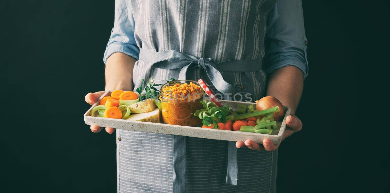 Crop anonymous female cook holding tray with assorted chopped products and jar with vegetable conservation on black background in studio