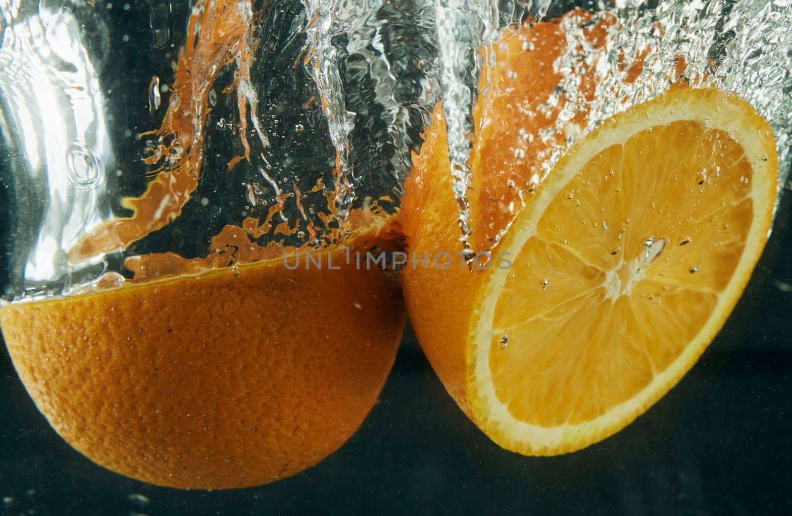 Halves of ripe tasty fresh unpeeled oranges falling in clear transparent water with bubbles in light studio on black background