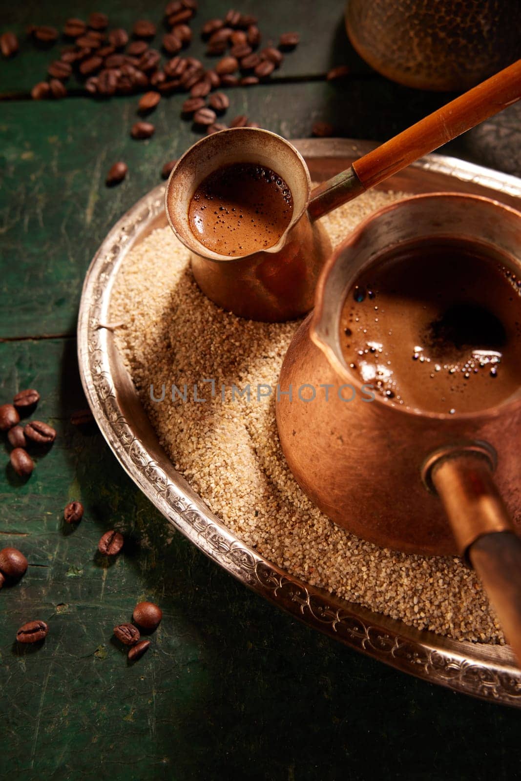 From above of metal cezves with freshly brewed Turkish coffee on tray with sand placed on table with scattered coffee beans