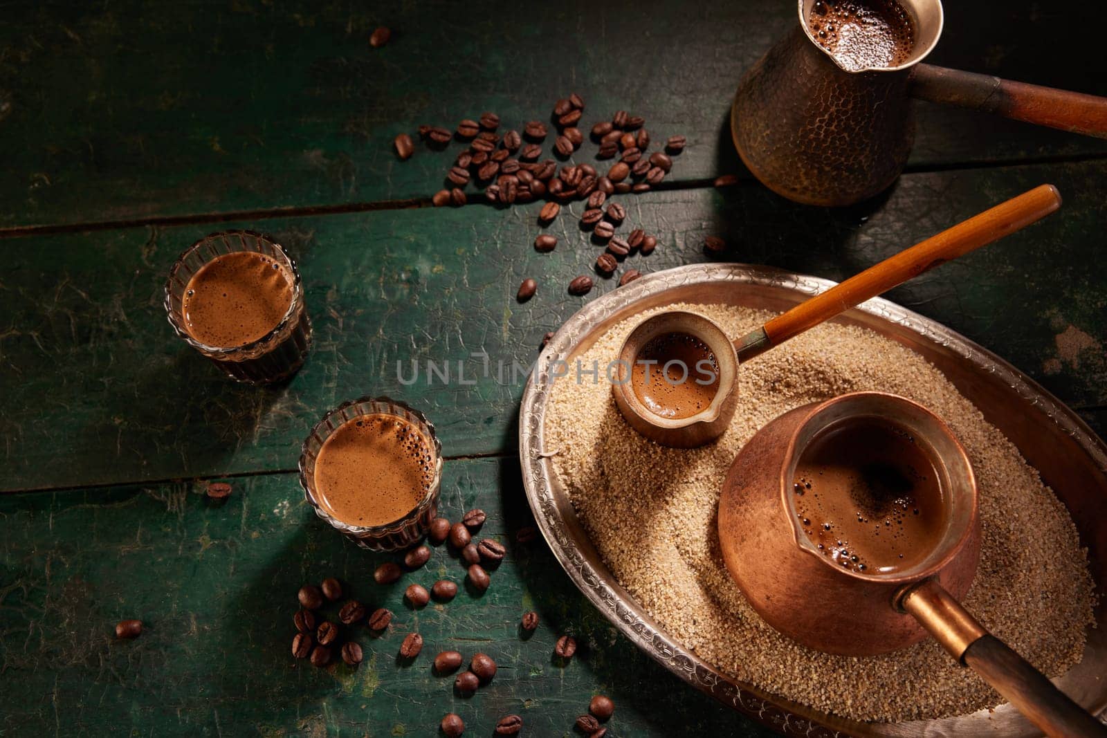From above of glasses and metal cezves filled with hot Turkish coffee placed on green table with sand and beans