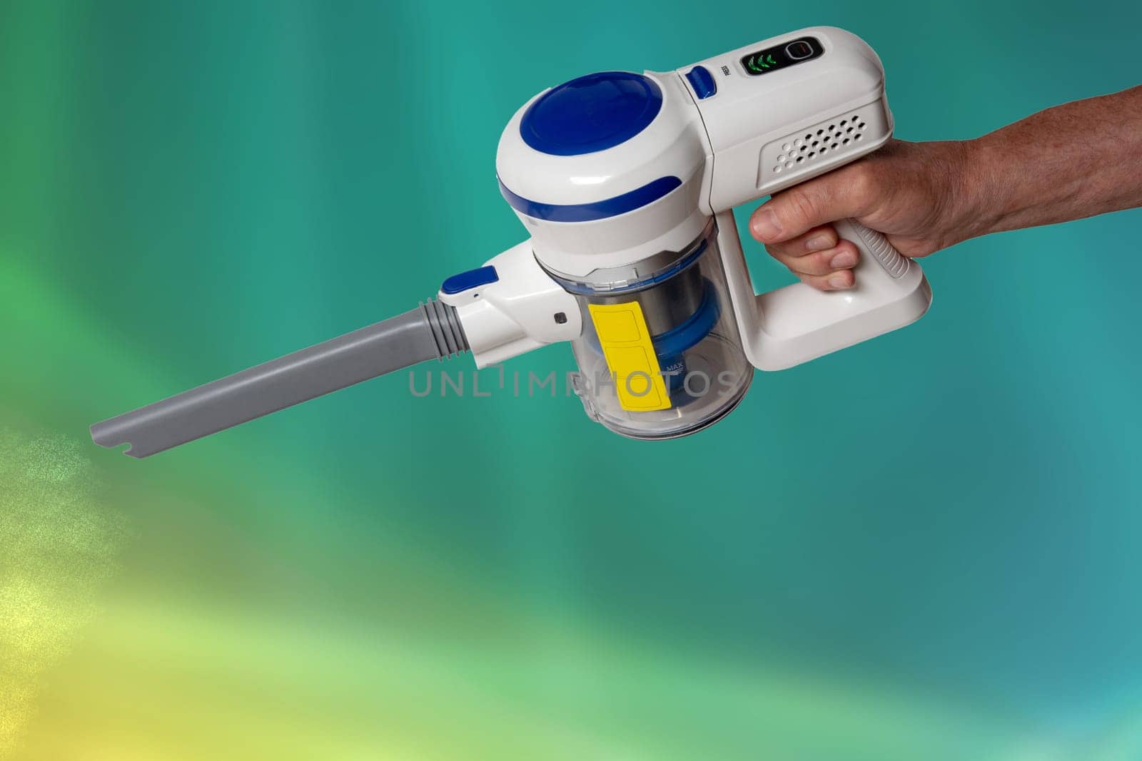 The male hand holds hold a modern cordless battery-powered vacuum cleaner over abstract blurred background. Space. Household cleanliness and hygiene.