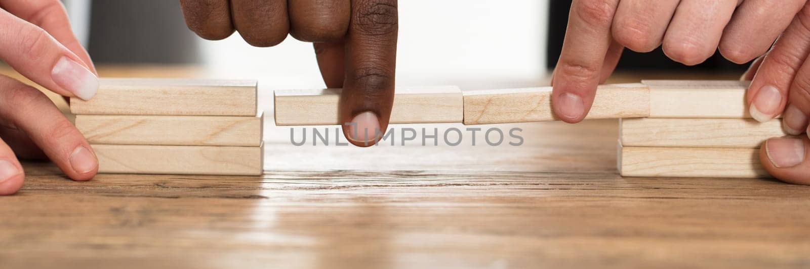 Businesspeople's Hand Holding Wooden Building Blocks To Form A Bridge Over A Gap On Table