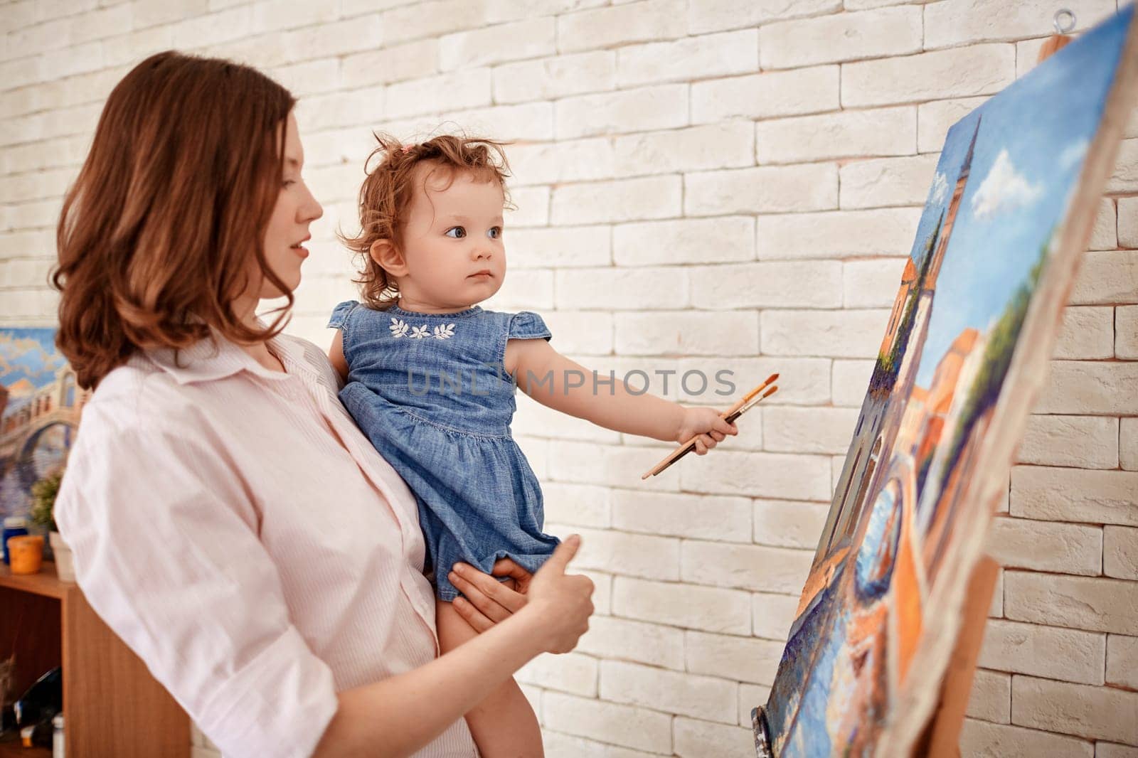 Young mother and her cute little daughter with paintbrushes looking at painting