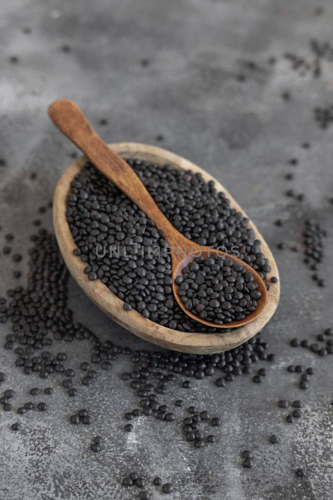 Bowl of dry black lentils beans with a spoon on grey table close up, copy space. Healthy eating, protein source for vegan and vegetarian diet. Traditional Asian and North African cuisines ingredient