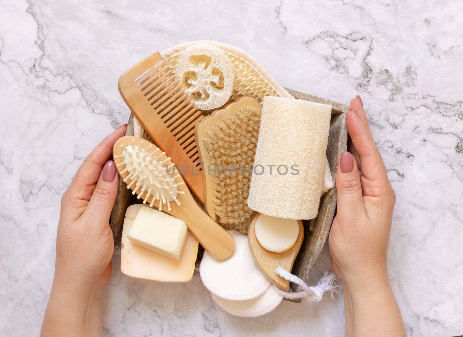 Hands holding tray of natural skin and hair care accessories over a marble table top view, copy space. Natural beauty products