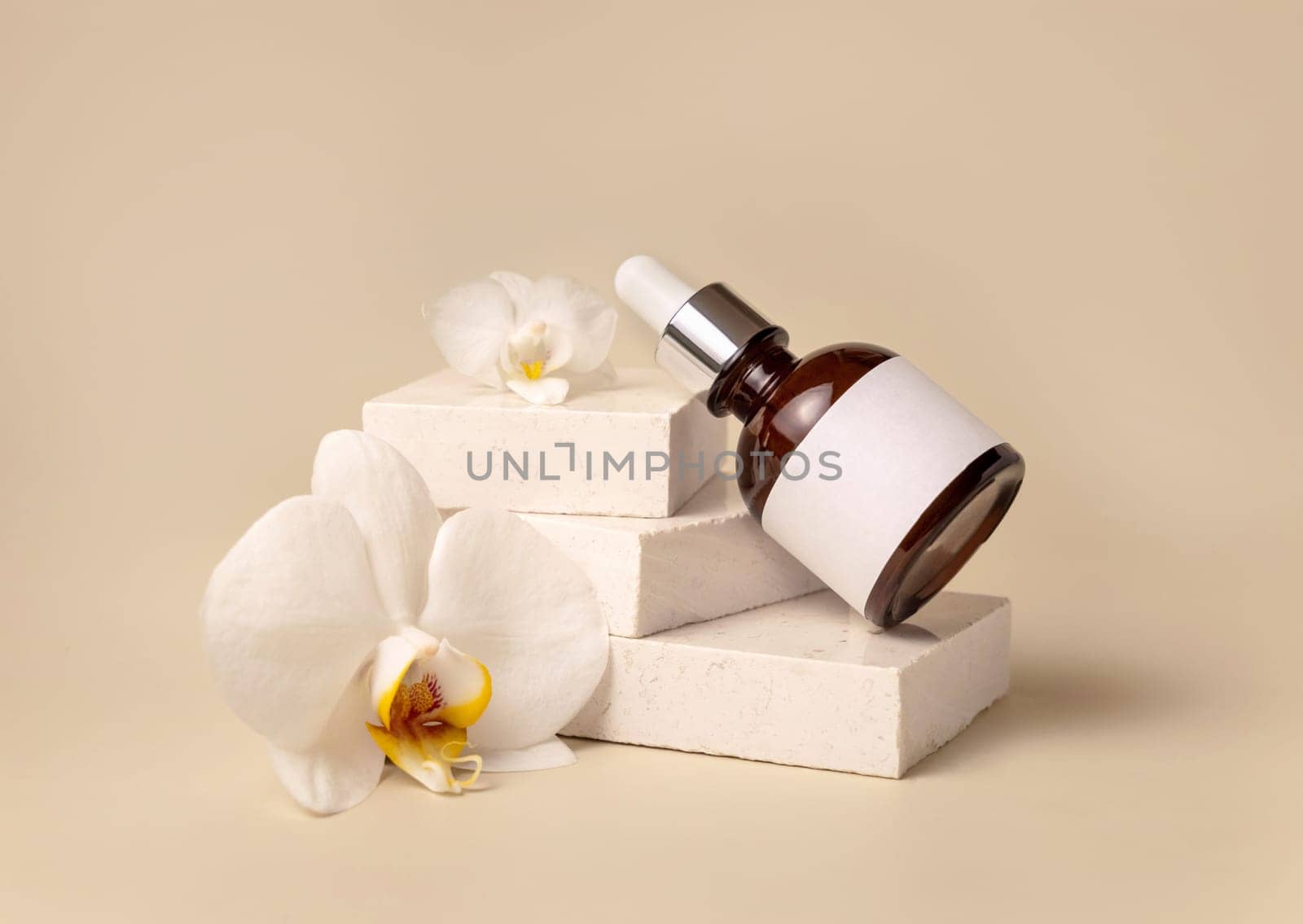 Brown glass dropper bottle on stone near white orchid flowers on light yellow, close up, mockup. Skincare handmade beauty product, serum or lotion. Exotic natural cosmetics..