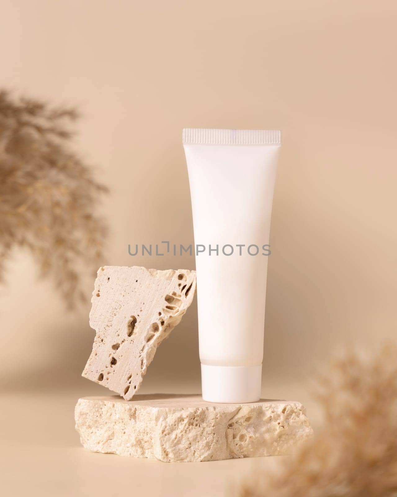 White plastic cream tube on travertine stone near dry pampas grass on light yellow, close up, mockup. Skincare beauty product. Natural cosmetics. Boho composition..