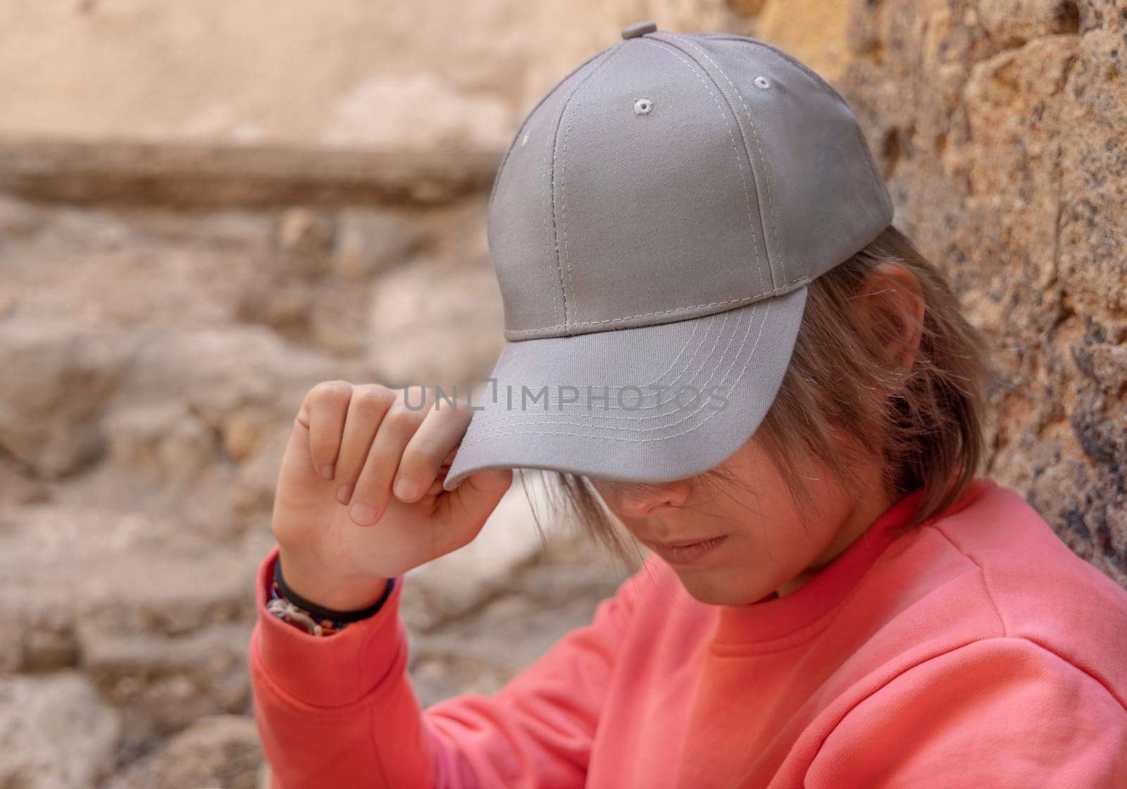 Teen girl in grey baseball cap and pink long sleeve shirt looking into a camera. Cap mockup. Serious Teenager girl near a stone wall outdoor touching the visor. Loneliness concept