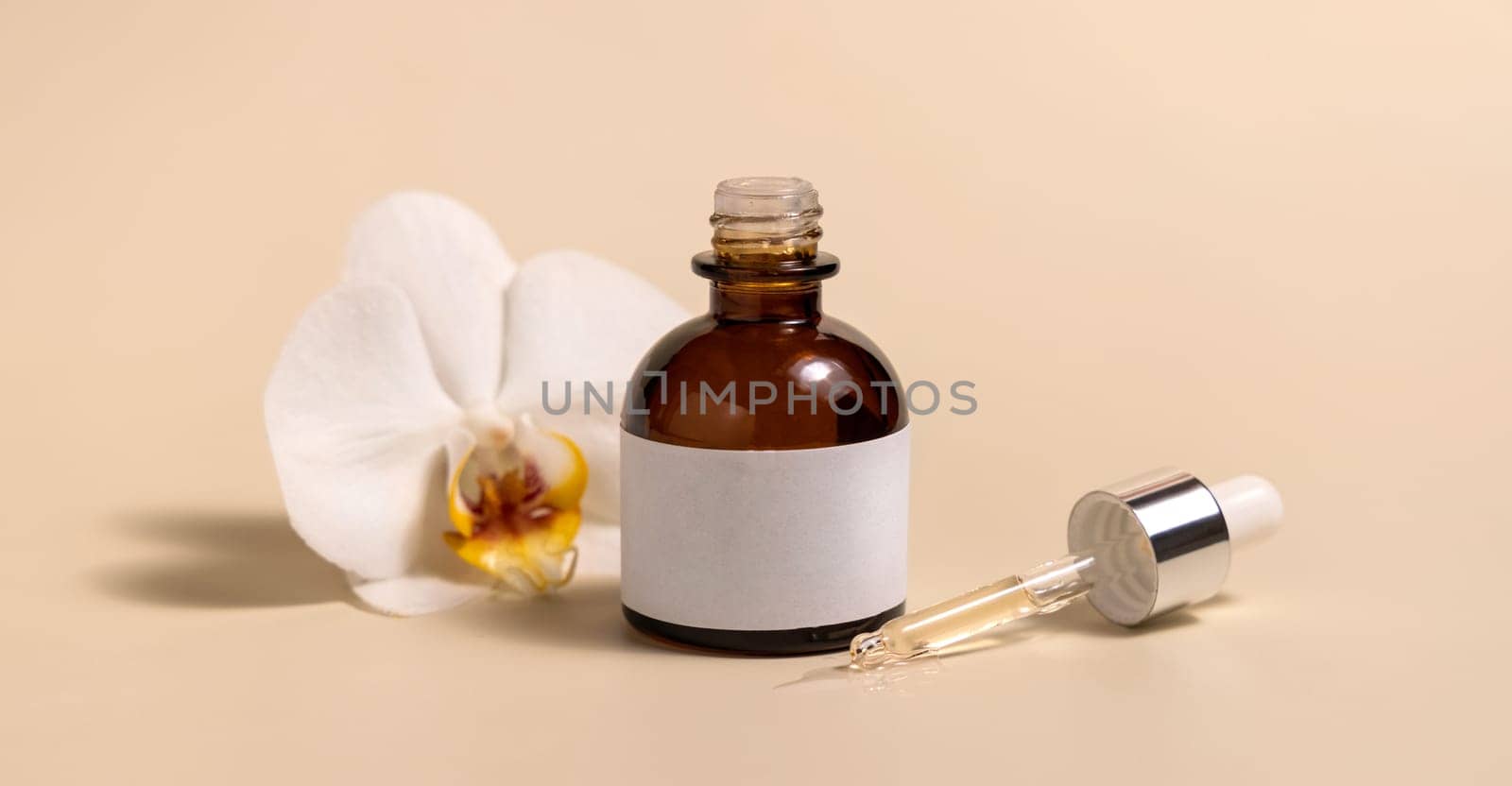 Brown glass dropper bottle near white orchid flowers on light yellow, close up, mockup. Skincare handmade beauty product, serum or lotion. Exotic natural cosmetics..