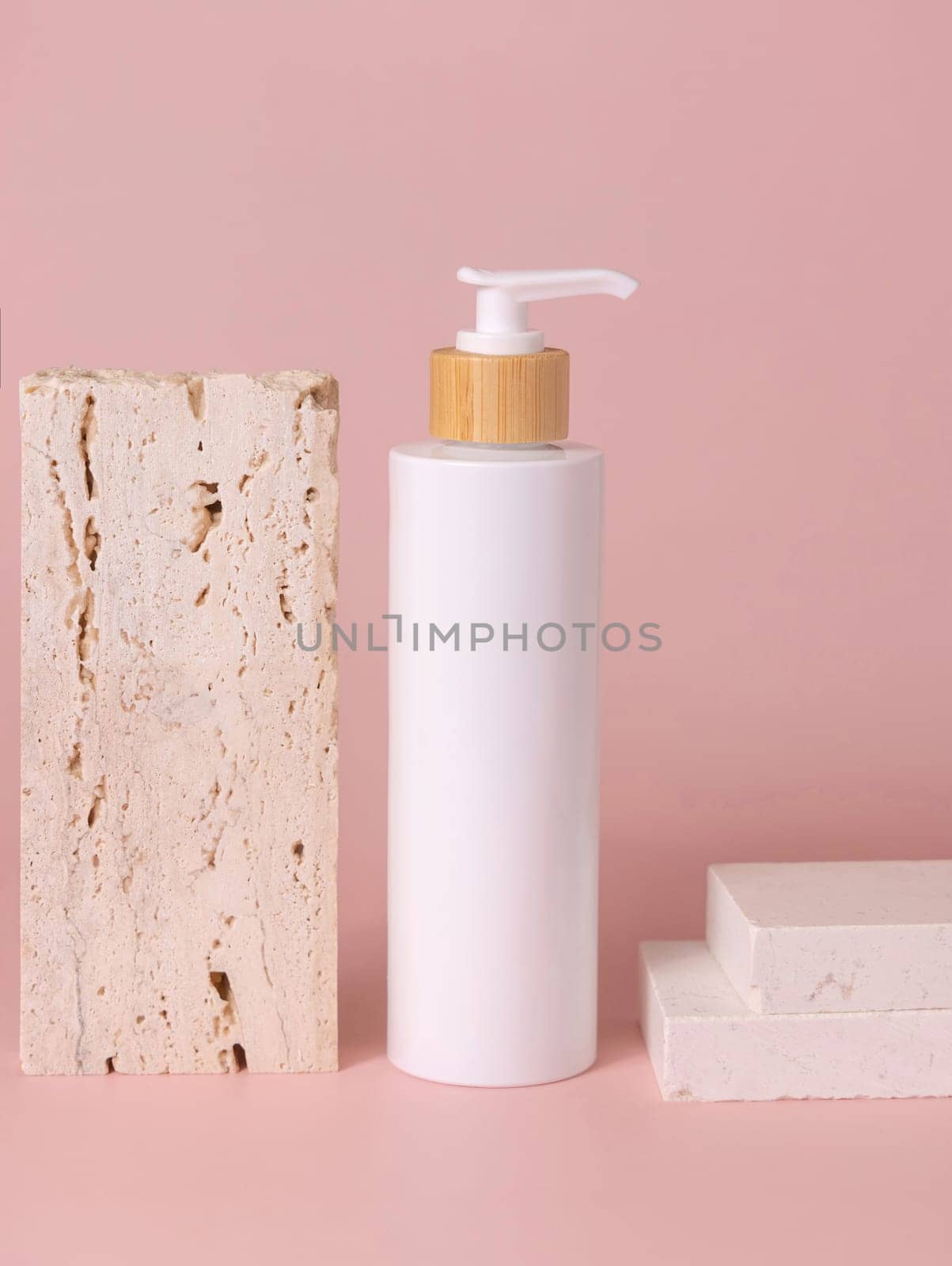 White one pump bottle near beige travertine stone on light pink, close up, mockup. Skincare beauty product. Natural cosmetics, lotion or cleanser. Minimal composition..