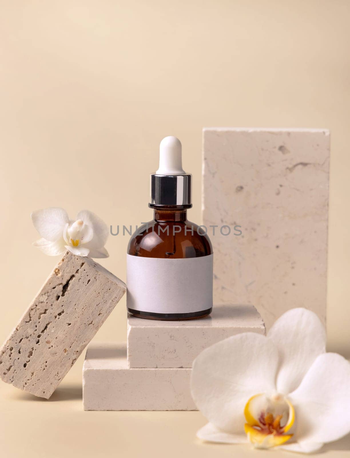 Brown glass dropper bottle on stone near white orchid flowers on light yellow, close up, mockup. Skincare handmade beauty product, serum or lotion. Exotic natural cosmetics..