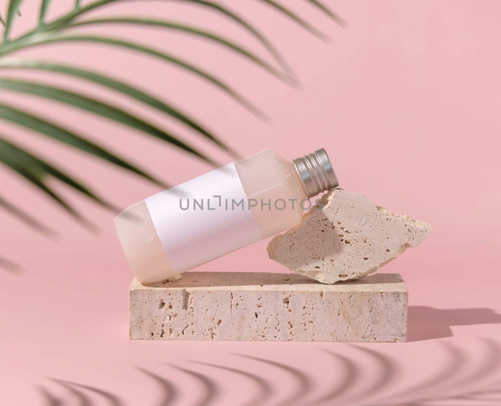 Cosmetic bottle with blank label lies on stone on light pink, palm leaf hard shadow , close up, mockup. Skincare beauty product. Natural cosmetics, lotion or cleanser..