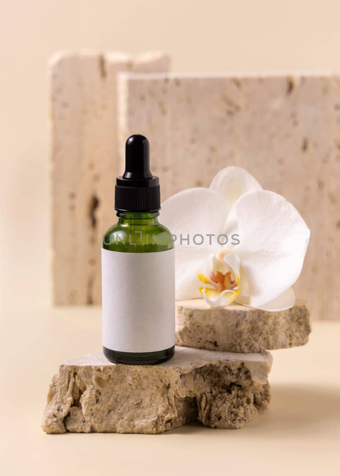 Green glass dropper bottle on stone near white orchid flower on light yellow, close up, mockup. Skincare handmade beauty product, serum or lotion. Exotic natural cosmetics..