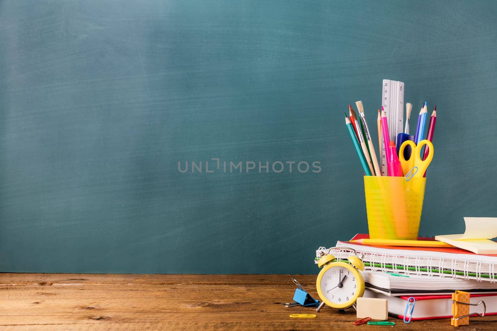 Many different various school, office supplies and stationery. Back to school concept. Notebook, book, pen, pencil, scissors, alarm clock, clock, sticker stand on a wooden table on a green background.