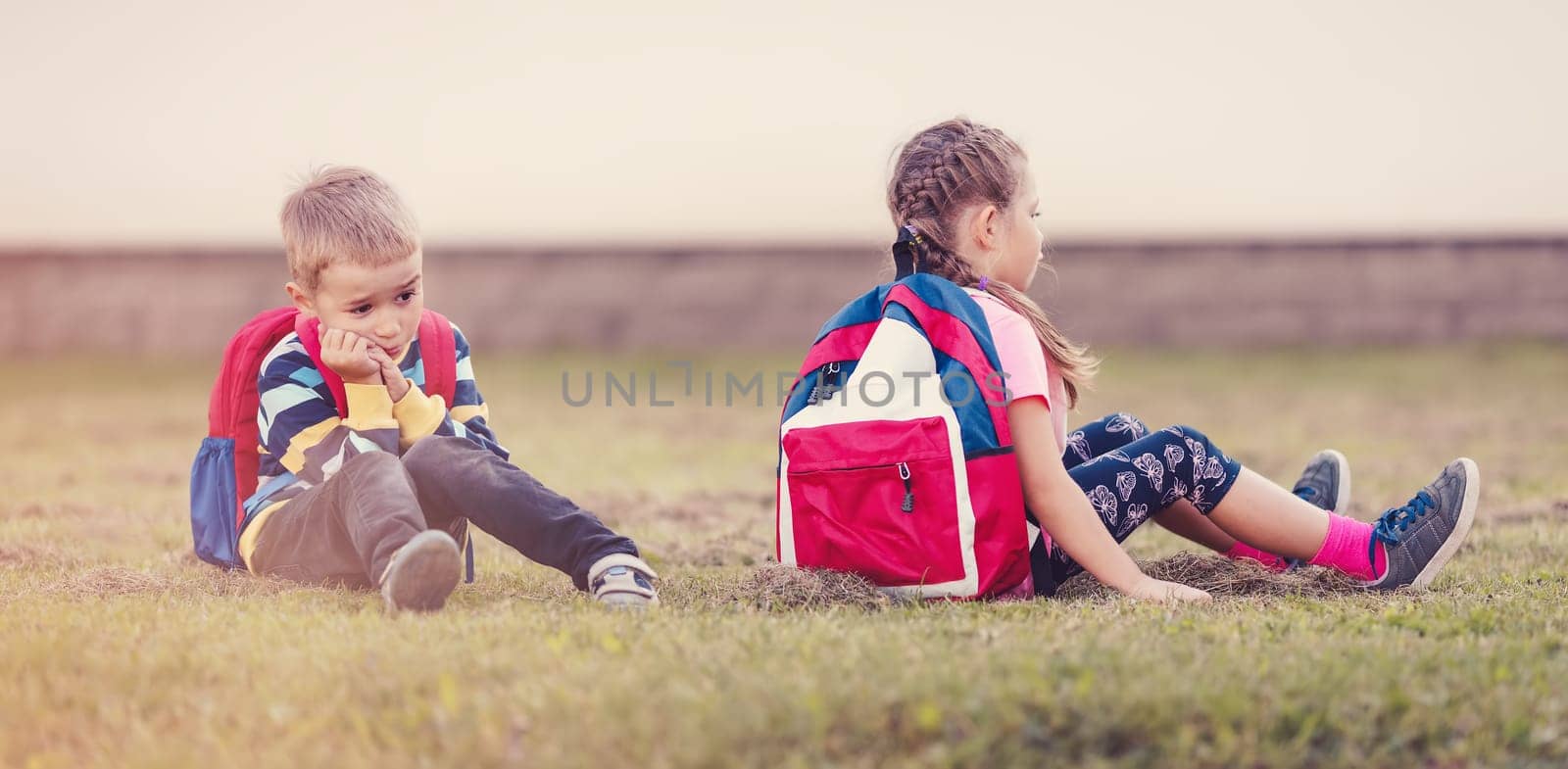 A boy and a girl quarreling and are sitting on the grass in the schoolyard back to back. Concept of friendship and back to school.