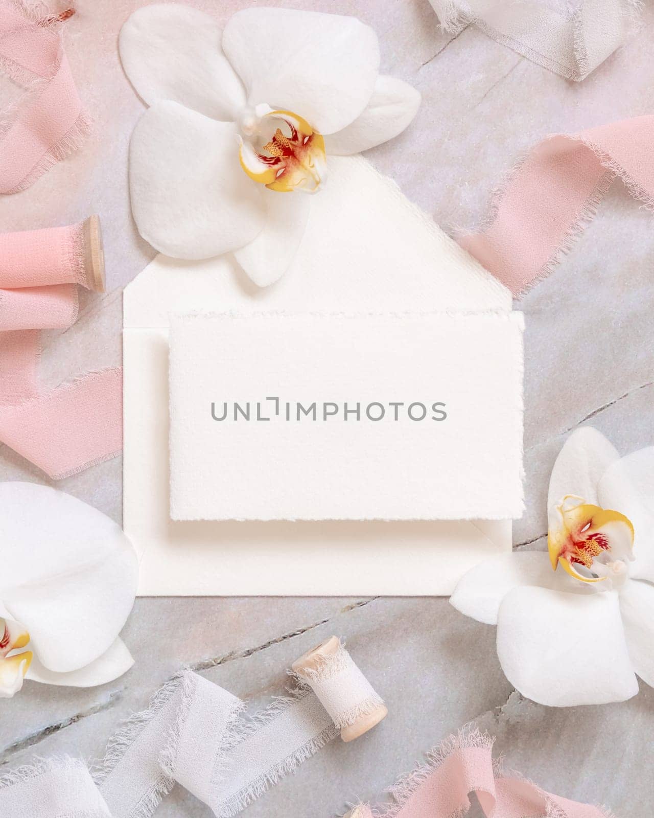 Wedding horizontal card and envelope near white orchid flowers and silk ribbons on marble top view, mockup. Romantic scene flat lay with handmade paper card