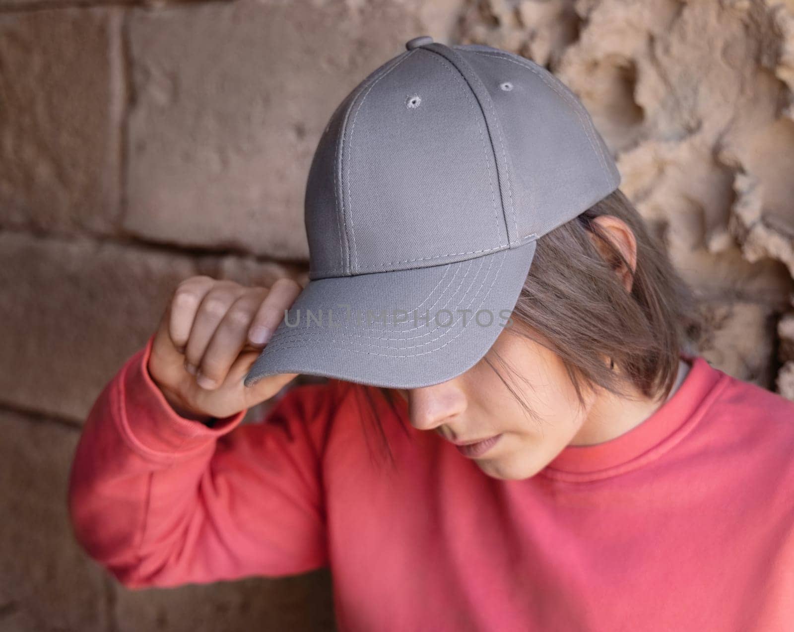 Teen girl in grey baseball cap and pink long sleeve shirt looking into a camera. Cap mockup. Serious Teenager girl near a stone wall outdoor touching the visor. Loneliness concept