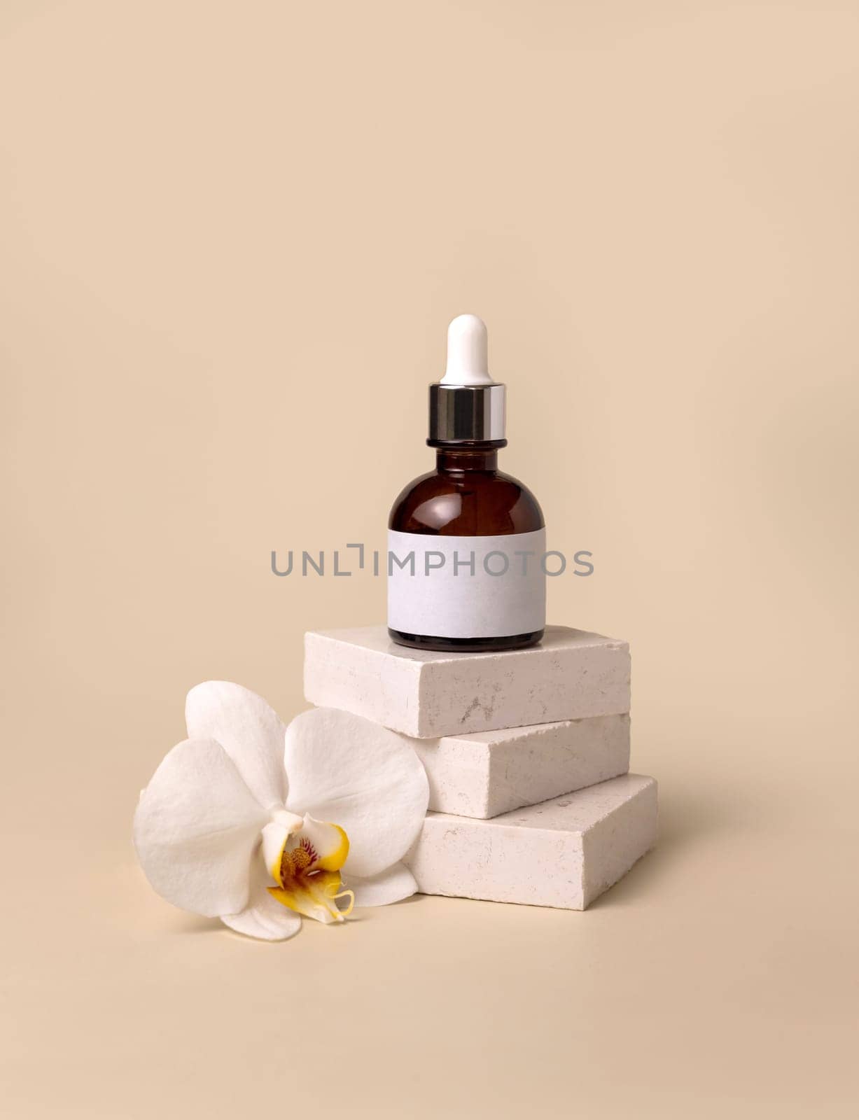 Brown glass dropper bottle on stone near white orchid flowers on light yellow, close up, mockup. Skincare handmade beauty product, serum or lotion. Exotic natural cosmetics..