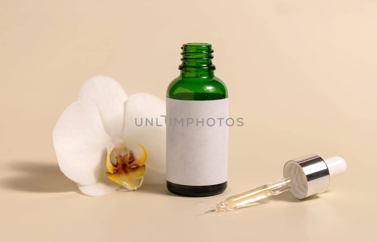 Green glass dropper bottle near white orchid flowers on light yellow, close up, mockup. Skincare handmade beauty product, serum or lotion. Exotic natural cosmetics..
