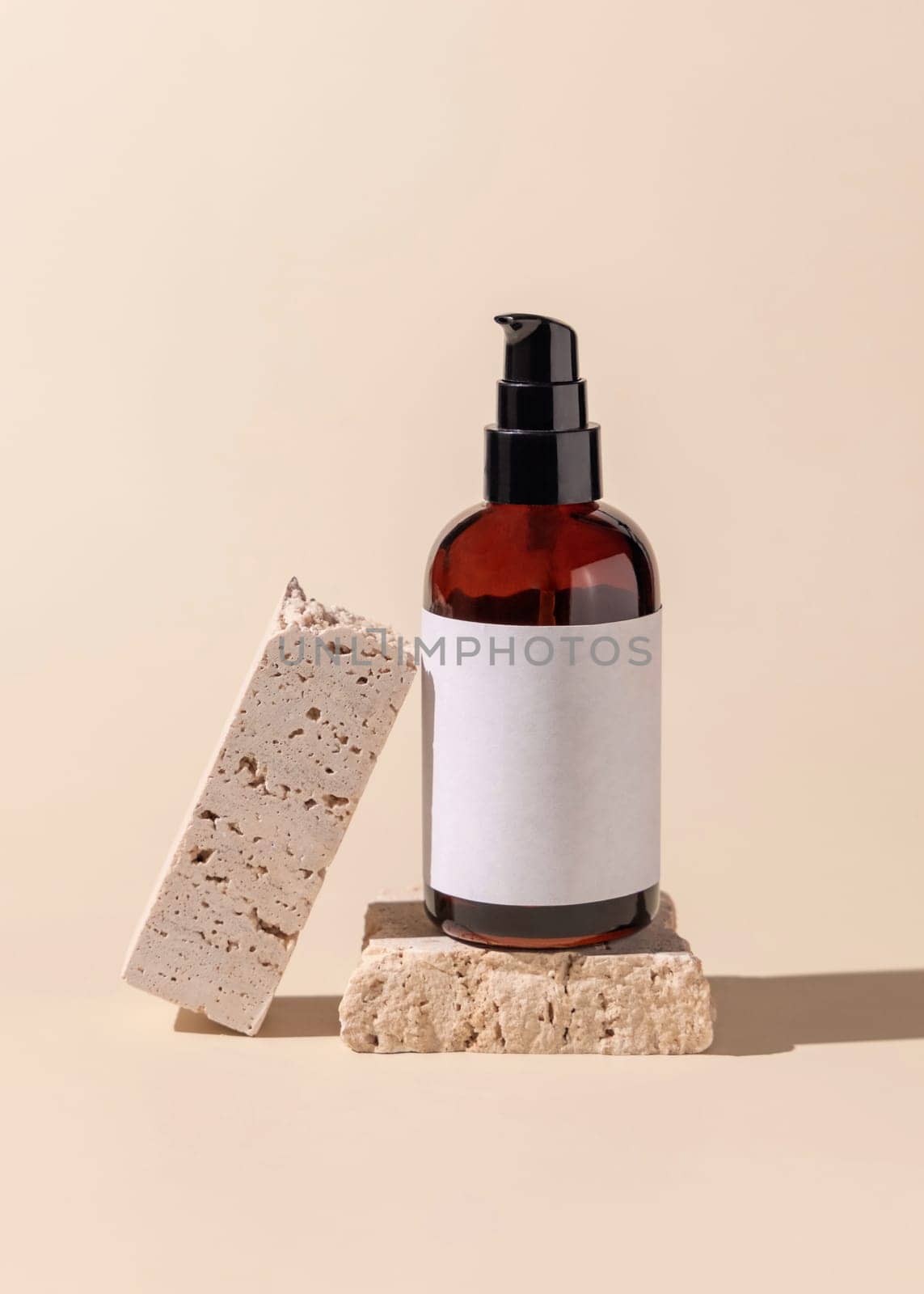 Brown glass one pump bottle near beige stones on light yellow close up, mockup. Skincare beauty product package. Natural cosmetics, minimal composition..