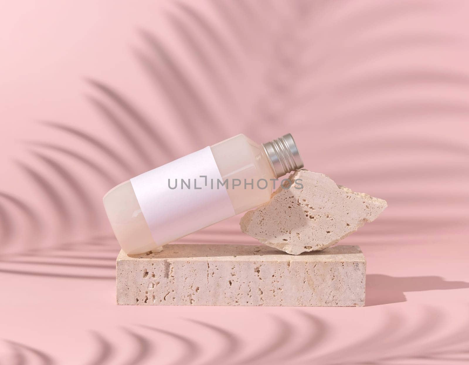 Cosmetic bottle with blank label lies on stone on light pink, palm leaf hard shadow behind, close up, mockup. Skincare beauty product. Natural cosmetics, lotion or cleanser..