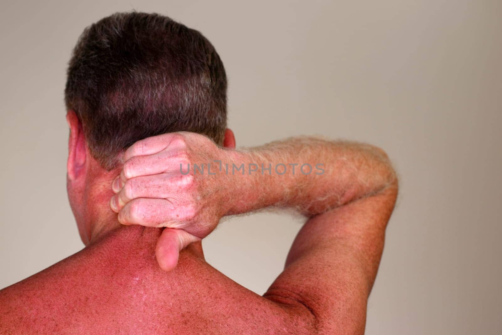 Mature adult white male seen from rear view massaging the back of his neck under his brown hair. Rear view of a naked caucasian man massaging his painful neck to help to relax his muscles close-up