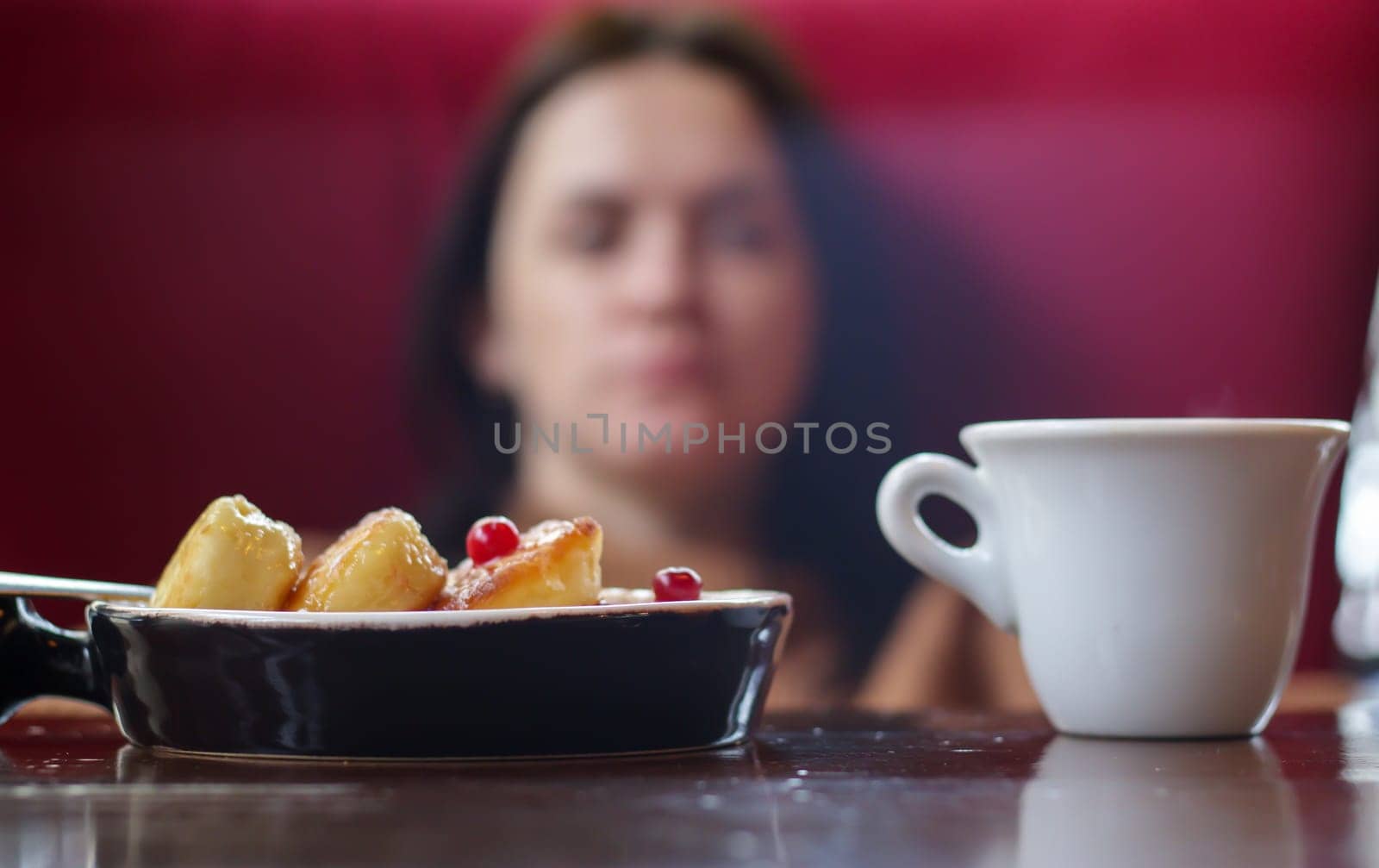 Portion of round paneer cheesecakes on a plate on a table with a white cup of coffee on a blurred background of an unrecognizable girl and restaurant interior. Healthy morning breakfast