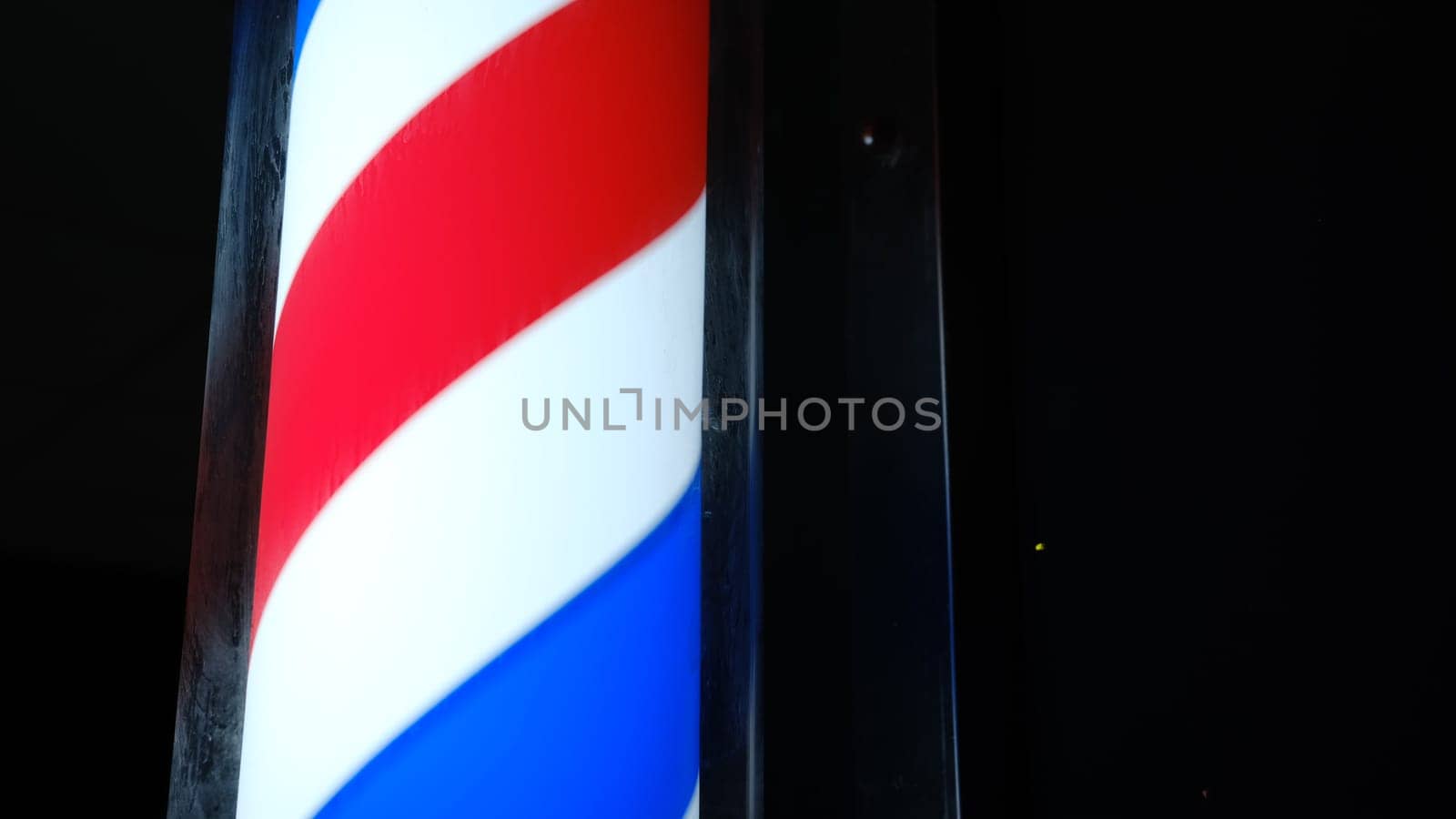 An old-styled barber shop sign, symbol. Round pole light in dark. Old-fashioned rotating led red white blue stripes pole light near men hair salon. Advertise of a small business outdoors. Hairdresser