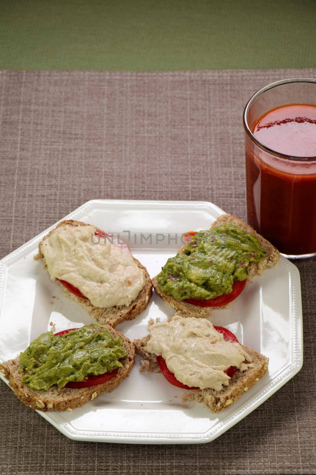 Four half slices of open faced sandwiches of hummus and tomato and guacamole and tomato on bread near a tomato vegetable beverage. Hummus, guacamole and tomato slices of four half slices of bread meal