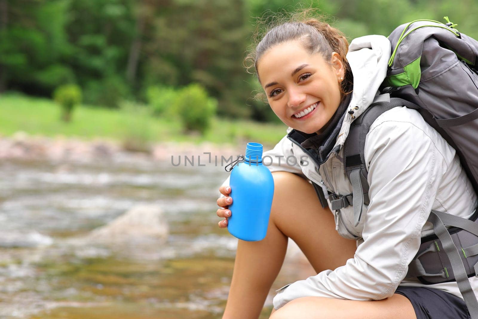 Happy hiker holding canteen looks at you in a riverside in the mountain