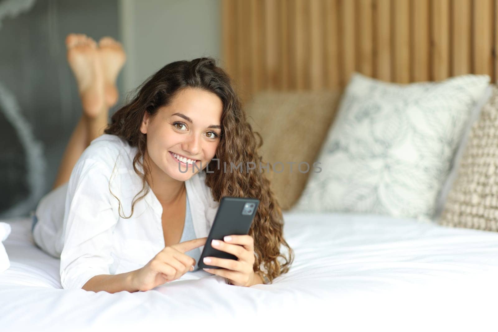 Happy woman holding smart phone looks at you lying on a bed