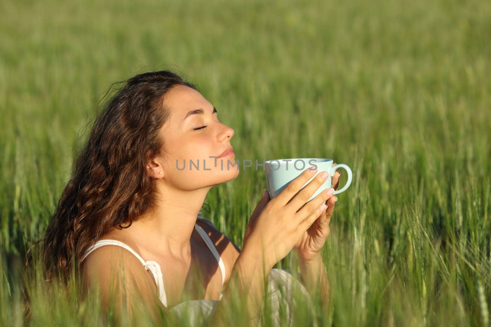 Woman relaxing smelling coffee from cup in a green wheat field