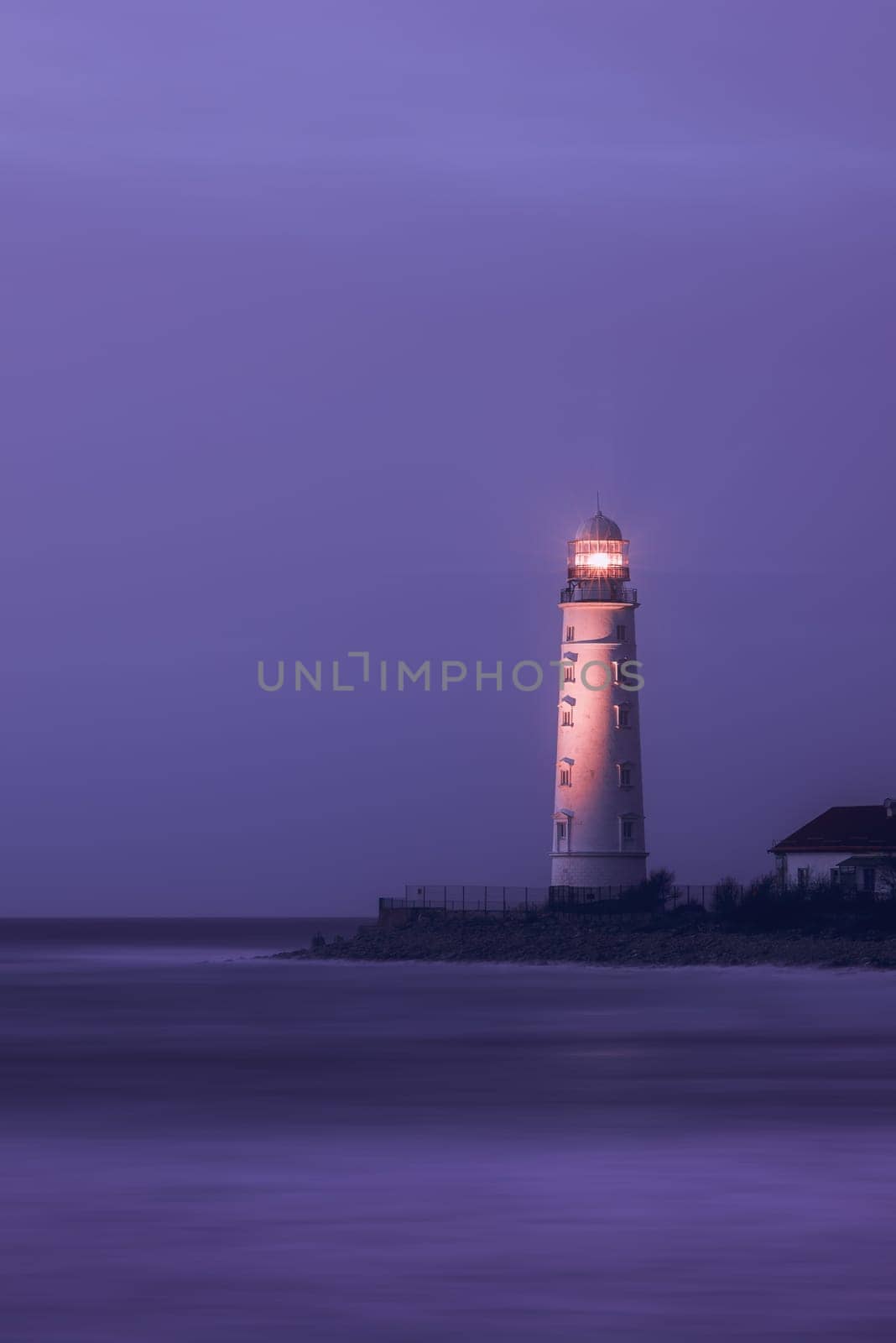 A lighthouse on the coast of the sea at dusk, its bright lamp is a landmark for ships. A lighthouse in the lilac twilight. Seascape, nature background.