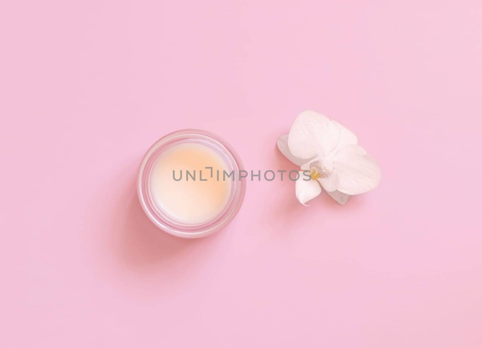 Small opened glass jar near white orchid flower on light pink top view, mockup. Skincare beauty product package. Exotic natural cosmetics, pastel minimal composition..