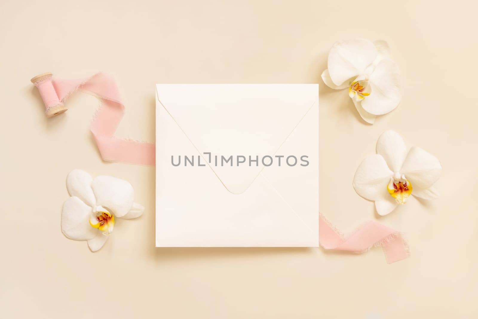 Wedding square envelope near white orchid flowers and silk ribbons on light yellow top view, mockup. Romantic scene with blank card flat lay. Valentines or Spring romantic concept