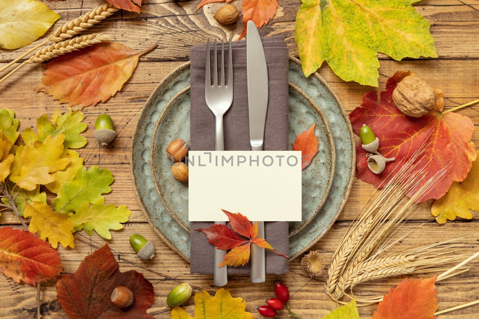 Autumn rustic table place with blank card between colorful leaves and berries, top view, mockup. Flat lay with horizontal card on wooden table, copy space. Thanksgiving or harvest concept