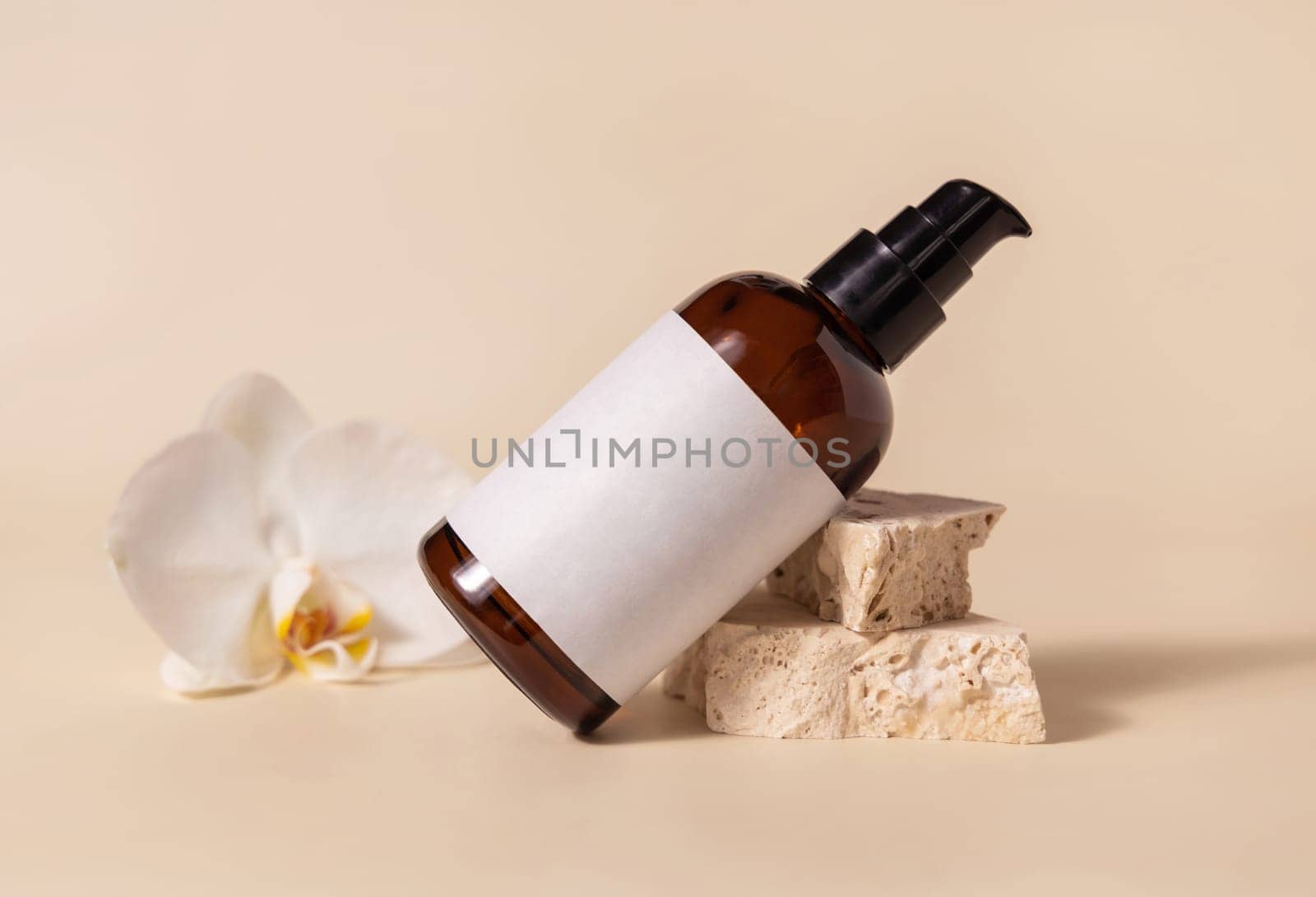 Brown glass one pump bottle near white orchid flower and beige stones on light yellow close up, mockup. Skincare beauty product package. Natural cosmetics, minimal composition..