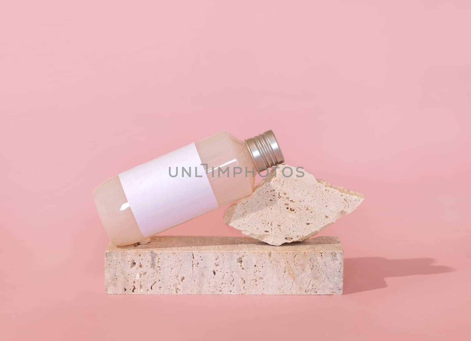 Bottle with blank label lies lies on travertine stone on light pink, close up, mockup. Skincare beauty product. Natural cosmetics, lotion or cleanser. Minimal composition..