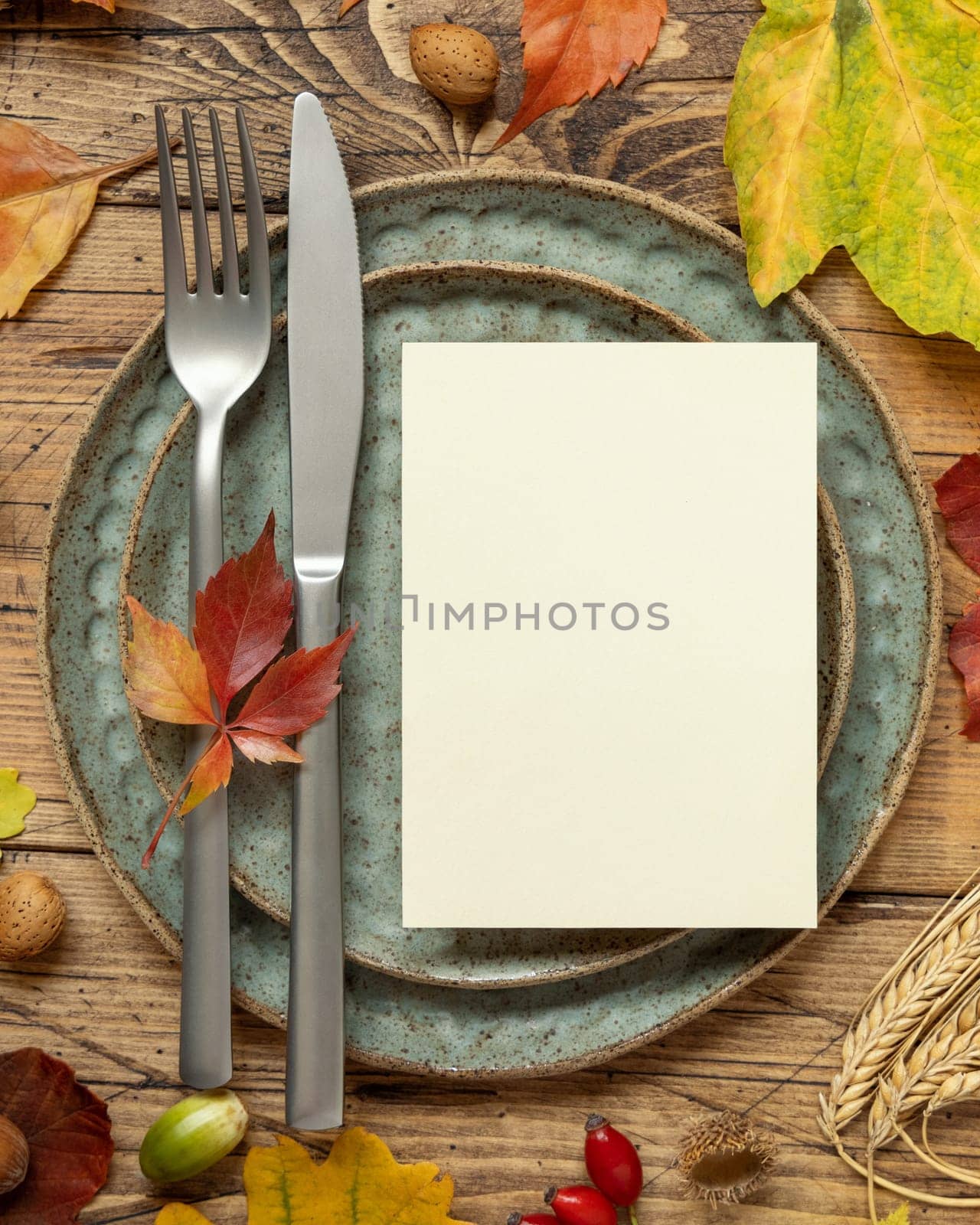 Autumn rustic table setting with blank card between colorful leaves and berries, top view. Vertical card mockup on wooden table, flat lay, copy space. Thanksgiving, halloween or harvest concept