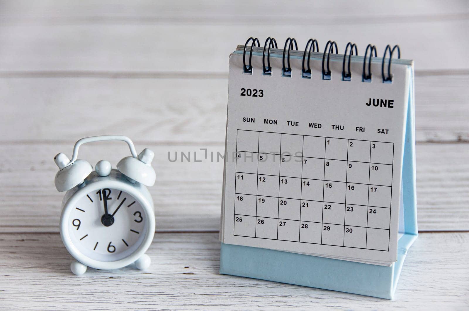 June 2023 white desk calendar on wooden table with alarm clock pointing at 12 o'clock.